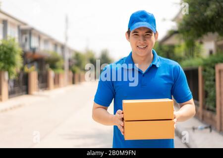 Asiatischer Auslieferungsjunge Mann in blauem, gleichmäßigem Lächeln und Kartonstapel im vorderen Hausdorf mit Kopfflächen. Werbung, Business, Tran Stockfoto