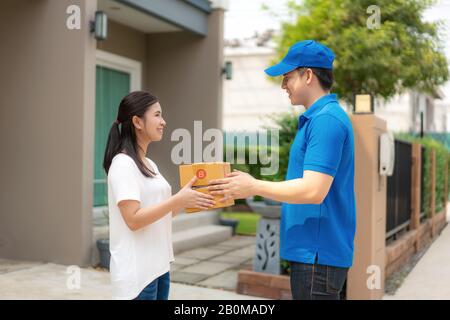 Asiatischer Auslieferungsjunge Mann in blauem, gleichmäßigem Lächeln und Kartonstapel im Vorderhaus und Asian Woman, die eine Lieferung von Kisten aus d annehmen Stockfoto