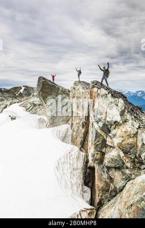 Drei Bergsteiger stehen auf einem felsigen Gipfel mit in der Luft erhobenen Armen Stockfoto