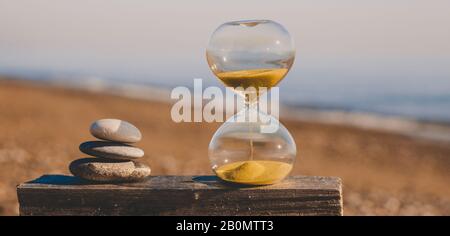 Sandglas auf einem Holzbrett mit drei Steinen in Form einer Pyramide, eine moderne Sanduhr an einem Strand mit goldenem Sand vor dem Hintergrund der n Stockfoto