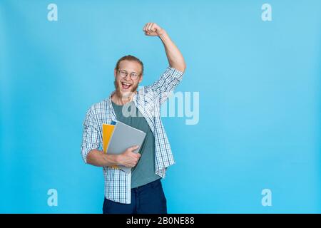 Bärtiger, glücklicher, aufgeregter Student in Brille hält eine Gewinn-Geste für das Copybook Stockfoto