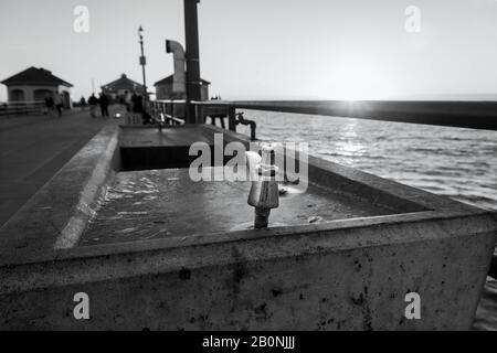 Nahaufnahme eines öffentlichen Trinkbrunnens aus Edelstahl und Beton am Huntington Beach Pier bei Sonnenuntergang. Sonnenstrahlen am Horizont. Stockfoto