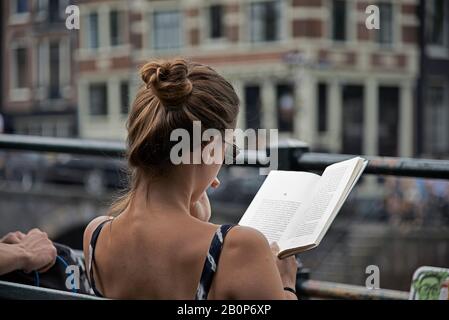 Junge kaukasische Frau, die auf einer Bank im Stadtzentrum von Amsterdam sitzt und bei Sommersonne ein Buch liest Stockfoto