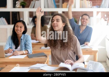 Lächelndes Mädchen der Teenager High School, das mit multinationalen Klassenkameraden im Klassenzimmer am Schreibtisch sitzt, Studenten der University College, die Hand heben, askin Stockfoto