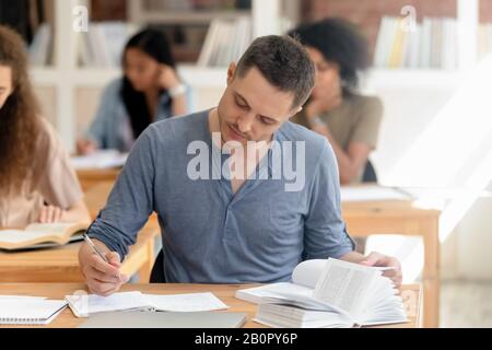 Attraktiver Teenager, der mit Lehrbuch im Klassenzimmer studiert, Notiz schreibt, am Schreibtisch sitzt, Studenten der College-Universität, die die Aufgabe vorbereiten, Finne passieren Stockfoto