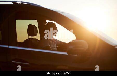 Silhouette eines jungen, ernsthaften Mannes, der in seinem Auto sitzt, Sonnenbrille trägt und den Sonnenuntergang betrachtet, helle Sonne auf dem Auto, Nahaufnahme, Seitenansicht Stockfoto