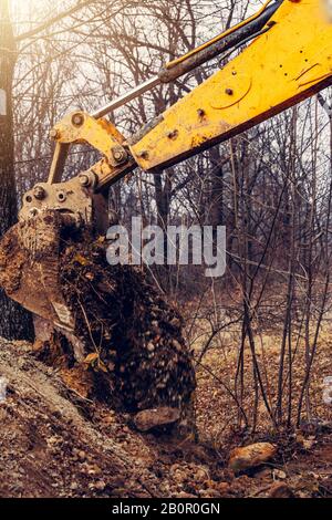 Ein gelber rostiger Industriebagger mit einem Eimer, der auf dem Boden liegt und an einer Baustelle in einem Teichwald den Boden gräbt. Stockfoto