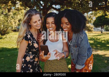 Portrait einer Gruppe von verschiedenen jungen Freundinnen, die etwas auf dem Smartphone im Park sehen Stockfoto