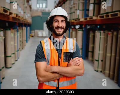 Porträt eines fröhlichen Vorgesetzten in einem Lager für die Lieferung und den Transport von Industriegütern mit weißem Helm und orangefarbener Weste Uniform stehend Stockfoto