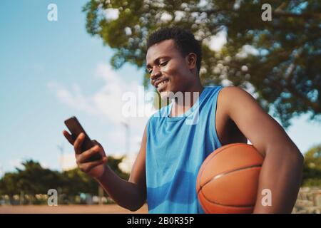 Porträt eines lächelnden jungen männlichen Basketballspielers, der den Ball in der Hand hält und Nachrichten auf seinem Smartphone im Park textet Stockfoto