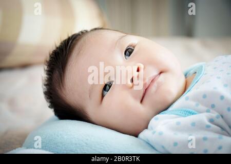 Asiatisches Babyportrait. Süßes asiatisches Mädchen öffnet die Augen auf dem komfortablen Bett, das Eltern am Tag mit einem kleinen Lächeln sucht. Fröhliches Kinderkonzept.