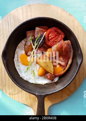 Frühstück in der Pfanne mit Spiegelei, Würstchen, Speck, Tomaten, Kartoffeln und Gemüse auf Holzblock Hintergrund auf dem Tisch in der Küche. Stockfoto