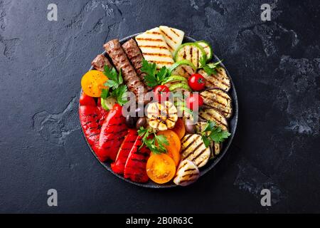 Gegrilltes Fleisch und Gemüse auf schwarzem Teller. Hintergrund aus schwarzem Stein. Draufsicht. Stockfoto