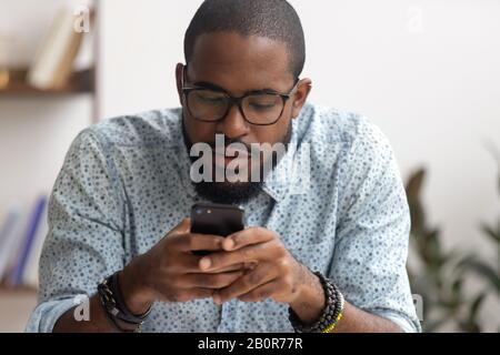 Fokussierter afroamerikanischer Geschäftsmann mit Smartphone im Büro, Blick auf Bildschirm, Chat im sozialen Netzwerk, seriöse Mitarbeiter lesen E-Mail, browsi Stockfoto