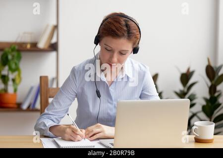 Fokussierte Frau in Kopfhörern mit Laptop, Notizen schreiben, Webinar zum Ansehen von Mitarbeitern, Schulung von Mitarbeitern, Sprache für Fernunterricht, E-Learning-Konzept, mA Stockfoto