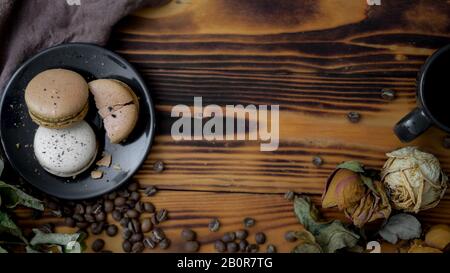 Draufsicht über Kaffee-Makronen auf schwarzem Teller mit Kopienraum, Kaffeebohne und Trockenrosen auf rustikalem Tischhintergrund Stockfoto