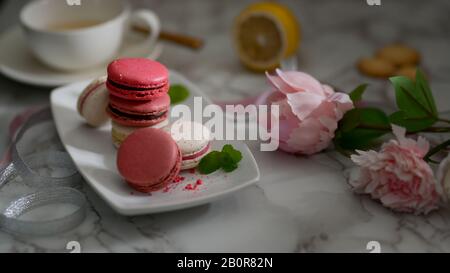 3/4-Schuss Farbenfroher Pastellmakarone auf weißem Keramikplatte mit Teetasse und Blume auf Marmorschreibtisch Stockfoto