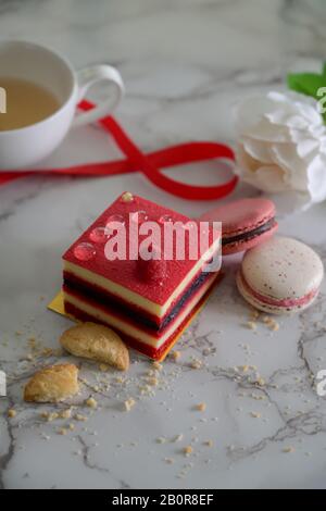 Zugeschnittenes Foto mit Himbeerkuchen und Makronen auf dem Marmorschreibtisch mit Teetasse, Keks und Blumen dekoriert Stockfoto