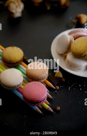 3/4-Schuss französischer farbenfroher Makaronen in weißer Platte und Bleistifte mit trockener Blume auf schwarzem Tisch Stockfoto