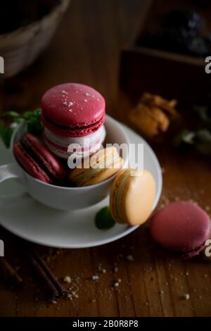 3/4-Schuss farbenfroher süßer Makronen in weißem Becher mit trockenen Rosen, die auf Holzschreibtisch dekoriert sind Stockfoto