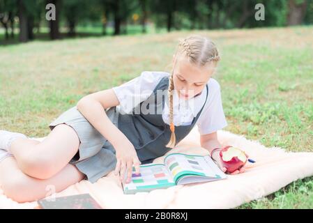 Schulmädchen liest Buch im Park über die Natur, kleines Mädchen isst Äpfel, Fernlehrgang Stockfoto