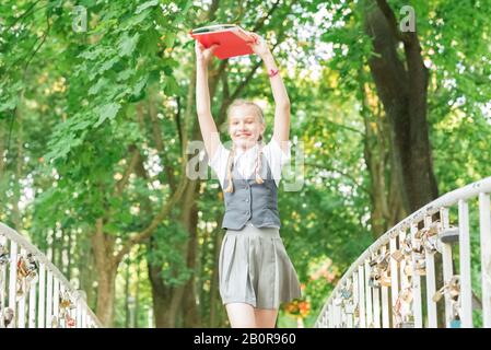 Student Schülerin mit Zöpfen in Uniform mit Bücher in die Hände über dem Kopf läuft glücklich über die Brücke im Sommer Park Stockfoto