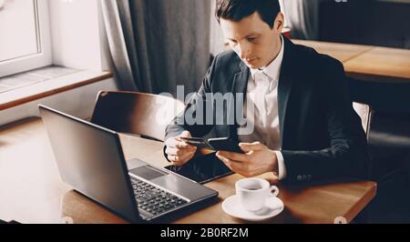 Kaukasischer Freiberufler mit schwarzem Haar, der einen Anzug trägt, stellt während einer Kaffeepause einige Daten einer Bankkarte in seinem Telefon vor Stockfoto