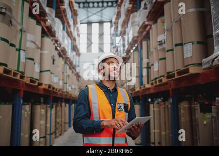 Niedriger Blickwinkel des jungen afrikaners, der eine reflektierende Jacke trägt, die das digitale Tablett im Fabriklager hält, lächelnd Stockfoto