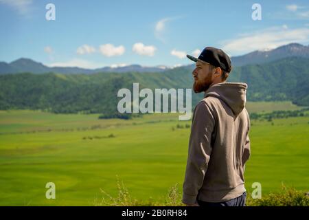 Ein rotbärtiger Mann im Hoodie, der vor grünem Feld und Berghintergrund steht. Sonniger Tag im altai-gebirge in sibirien Stockfoto