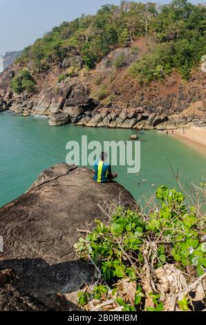 Der Junge sitzt auf einem Felsen, der in der Aussicht auf den Schmetterlingsstrand, Canacona, Goa, Indien einbricht Stockfoto