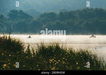 Fischermänner fischen auf einem Boot. Frühmorgens Frühlingslandschaft mit Fluss im Vordergrund und einem grünen Ufer mit Büschen und Bäumen im Hintergrund Stockfoto