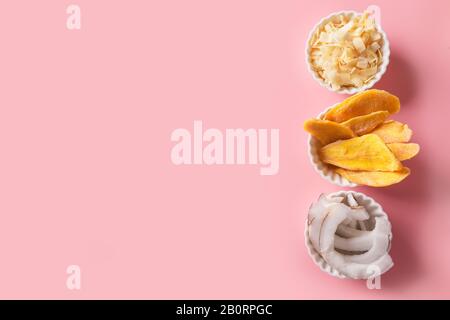 Dehydrierte und getrocknete Mango- und Kokosnuss-Chips in weißer Schüssel auf Rosa. Ansicht von oben. Leerzeichen für Text. Leckere und gesunde vegane Süßigkeiten. Stockfoto