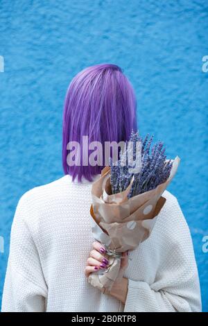 Junge Dame mit violettem Haar, weiße Bluse, stehend, mit Lavendelbouquet in Papierverpackung in der rechten Hand vor der blauen Wandrückengrou zurückdrehen Stockfoto