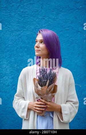 Portrait eines Mädchens mit violettem, farbenfrohem Haar, in weißer Strickjacke und blauem Rock, das einen Lavendelstrauß in Papierhüllen in ihren Händen hält. Mädchen steht Stockfoto