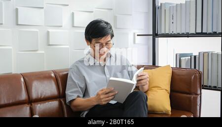 Senior Asian man liest Buch auf dem Sofa im Wohnzimmer zu Hause, Portrait of Asian Altern man ist Entspannend und Glücklich Mit Read a Magazine Stockfoto