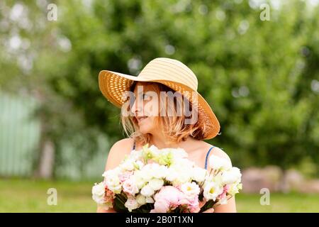 Eine junge Frau hält einen großen Blumenstrauß an Ponys vor sich. In einem Strohhut mit breiter Krempe. Blickt nach rechts. Sonniger Sommertag. Stockfoto