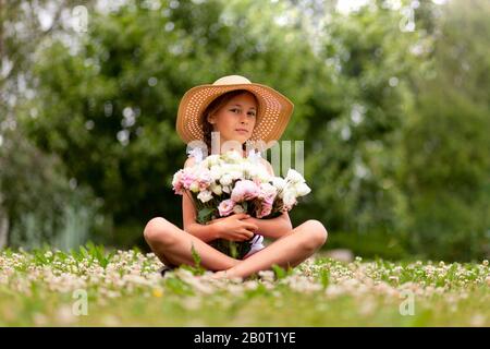 Mädchen, die einen großen Blumenstrauß an Pfingstrosen halten. Der junge Teenager sitzt in einem Hut mit breitem Rand auf einer blühenden Wiese. Mädchen mit zwei Zitzen. Sonniger Sommertag. Stockfoto