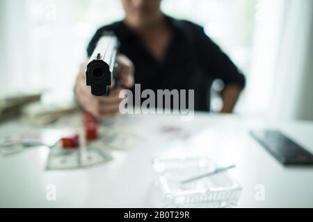 Junger Mann, der auf einem Tisch sitzt und eine Waffe hält, hat Alkoholglas und Geld auf dem Tisch. Stockfoto