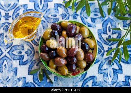 Auswahl an frischen Oliven auf einem Teller mit Olivenbäumen. Blauer Textilhintergrund. Draufsicht. Stockfoto