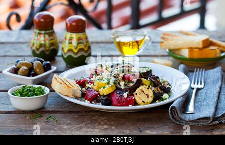 Gegrillter Gemüsesalat mit Fetakäse in weißer Platte auf Holztisch. Hintergrund im Freien. Stockfoto
