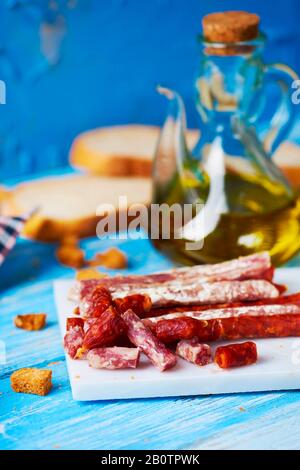Einige Stücke Fuet und Chorizo, zwei verschiedene spanische Wurstwaren, auf einem weißen Teller auf einem blauen Holztisch, neben einigen Scheiben Brot an Stockfoto