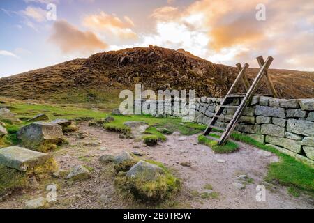 Mourn Wall mit Leiter auf der Hares Gap, die den Berg Slieve Bearnagh mit Sonnenuntergang zu goldener Stunde beaufsichtigt. Mourne Mountains Range in Nordirland Stockfoto