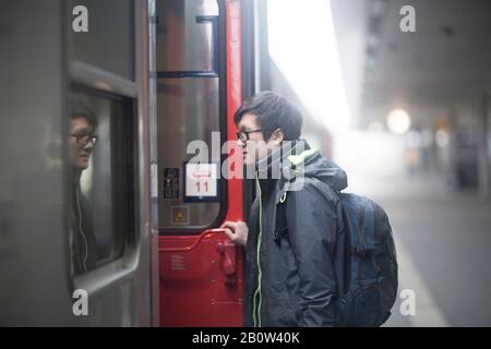 Junger Asiat, der auf dem Bahnsteig eine Brille trägt und in den Personenzug einfährt. Stockfoto