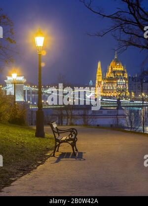 Budapest, Ungarn - Bank- und Lampenposten in einem Park im Bezirk Buda mit dem Parlament Ungarns und der Szechenyi-Kettenbrücke im Hintergrund zur Winterzeit Stockfoto