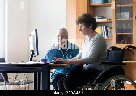 Ältere Frau und Frau im Rollstuhl sitzen an einem Tisch und arbeiten am Computer. Stockfoto