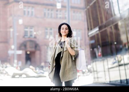 Porträt der jungen Frau mit langen braunen Haaren mit beigefarbenem Mantel, Einkaufstasche und Blick auf die Kamera. Stockfoto