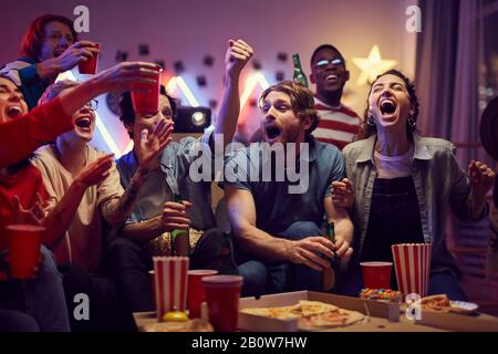 Eine Gruppe junger Fans, die auf dem Sofa sitzen und Bier trinken und Pizza essen, während sie zu Hause Fußballspiele auf dem Fernseher sehen Stockfoto