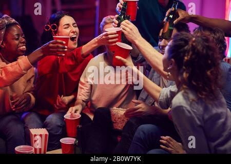 Eine Gruppe glücklicher Freunde, die mit Alkoholgetränken toben, während sie auf dem Sofa sitzen und ihr Treffen feiern Stockfoto