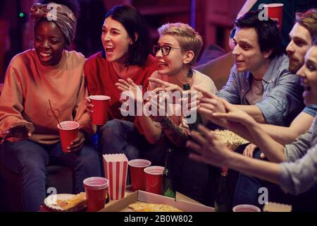 Junge Leute trinken Bier und essen Popcorn, während sie auf dem Sofa sitzen und gemeinsam auf der Party Fernsehen Stockfoto