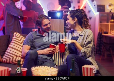 Junges Paar sitzt auf dem Sofa und trinkt Bier und spricht sich während der Heimparty bei ihren Freunden an Stockfoto